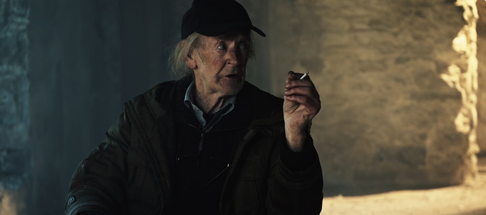 Production still — man in a cap lighting a cigarette in a stone interior