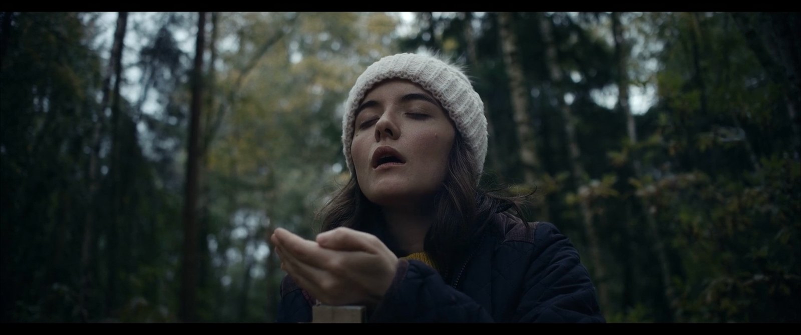 Production still — woman in a forest in overcast daylight