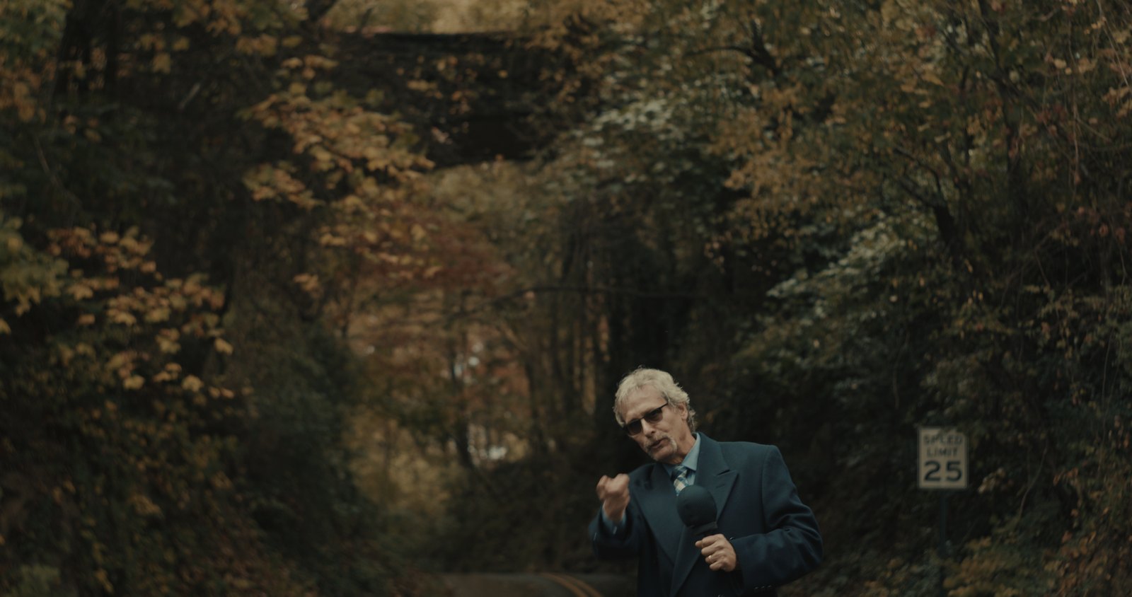 Production still — man in a suit on a wooded road in autumn