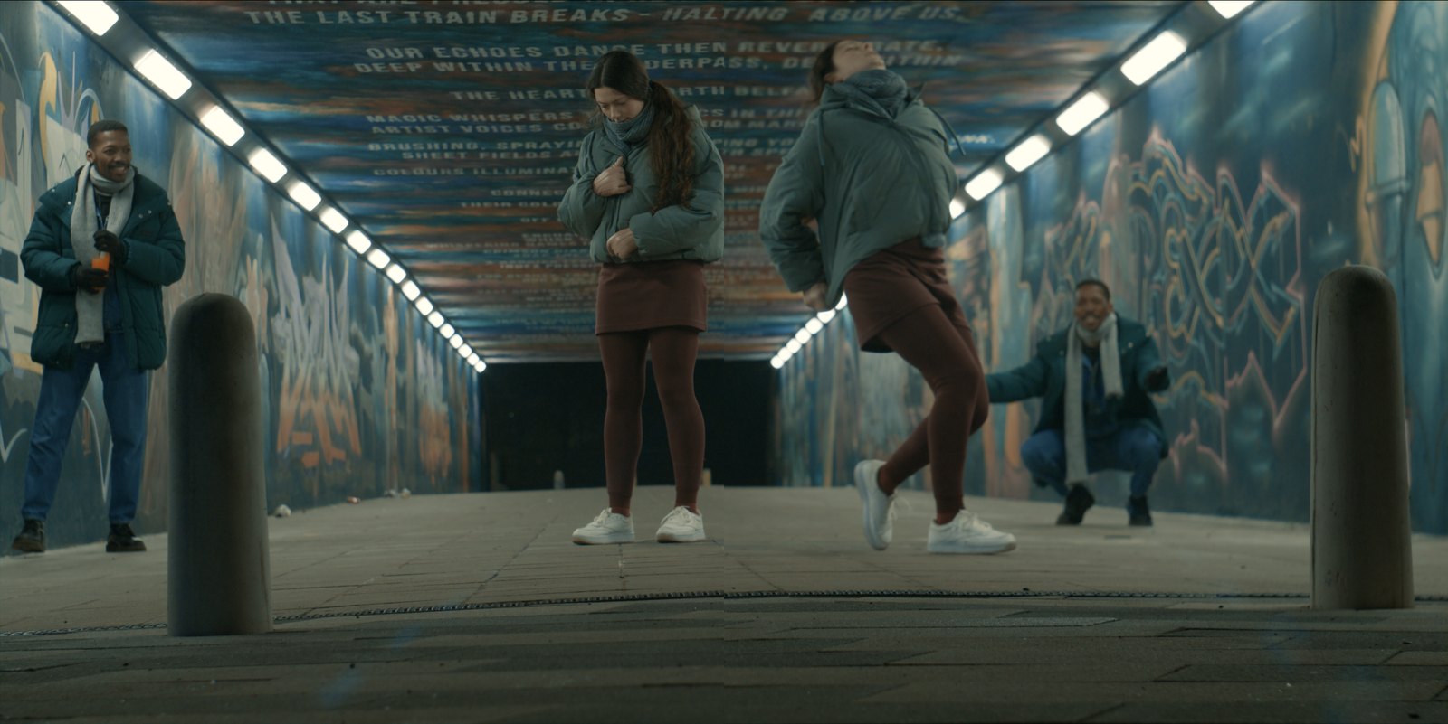 Production still — figures in a mural-painted pedestrian underpass